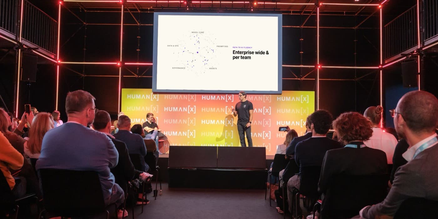 kju.ai pitching in the Human[X] Europe startup competition final in Amsterdam, with the slide 'Path to AI fluency: enterprise wide and per team' on the big screen and a full audience in front of the stage
