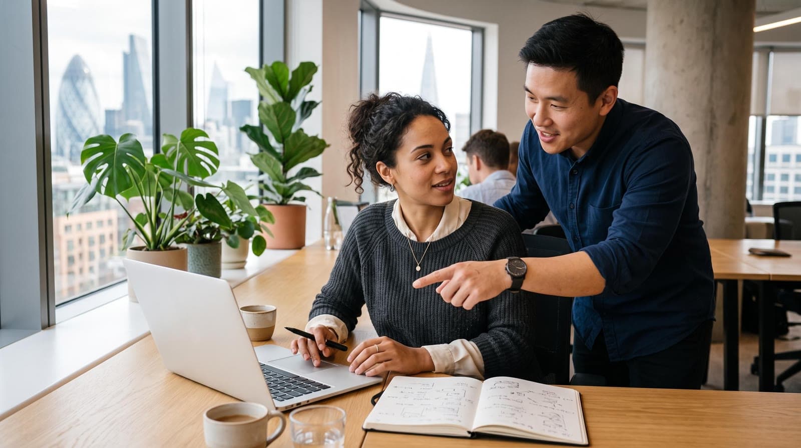 Two colleagues working through a problem together at a desk in a bright office, one taking handwritten notes beside an open laptop, illustrating AI literacy as ongoing practice rather than a one-off course
